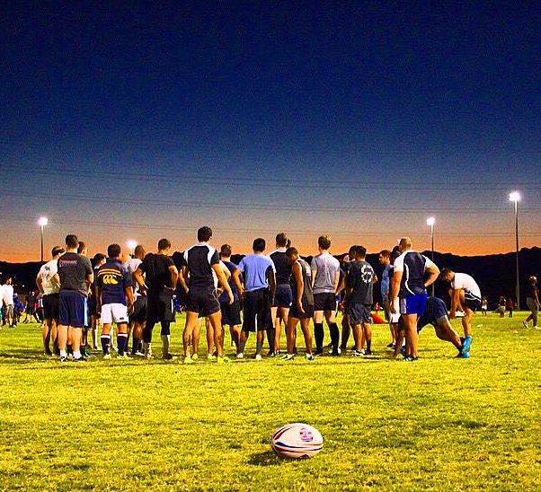 Downtown Rugby players gathered on a floodlit field at dusk for a summer touch session, rugby ball in the foreground